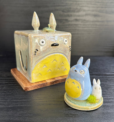 Set of ceramic sculptures, one cube shaped and modeled to look like Totoro. Nearby are a small blue and white chibi Totoro, standing on a small circle platform with a grass tuft.