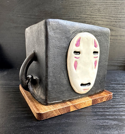 Cube shaped ceramic sculpture, painted black and with a face mask of No Face from Spirited Away. An arm comes out of its side and lifts up its cloak.