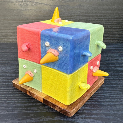 Ceramic cube made up of 8 different colored cubes: green, yellow, red and blue. Bird beaks come out of one side and little wings come out of other sides.