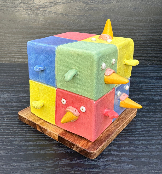Ceramic cube made up of 8 different colored cubes: green, yellow, red and blue. Bird beaks come out of one side and little wings come out of other sides.