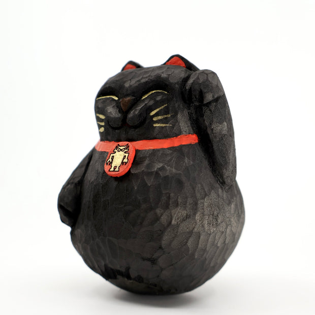 A rounded whittled wooden sculpture of a black maneki cats, smiling with closed eyes and one paw raised. It has gold whiskers and eyelids and wears a red collar with a gold Big Boss Robot on it.