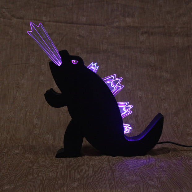 Die cut wooden silhouette of Godzilla, made into a lamp. A beam of light comes out of its mouth and it has lit up spikes along its back.