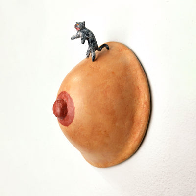 Sculpture of a single boob with a painted nipple. A small sculpted gray tabby cat stands atop it on its hind legs, with a fierce expression.