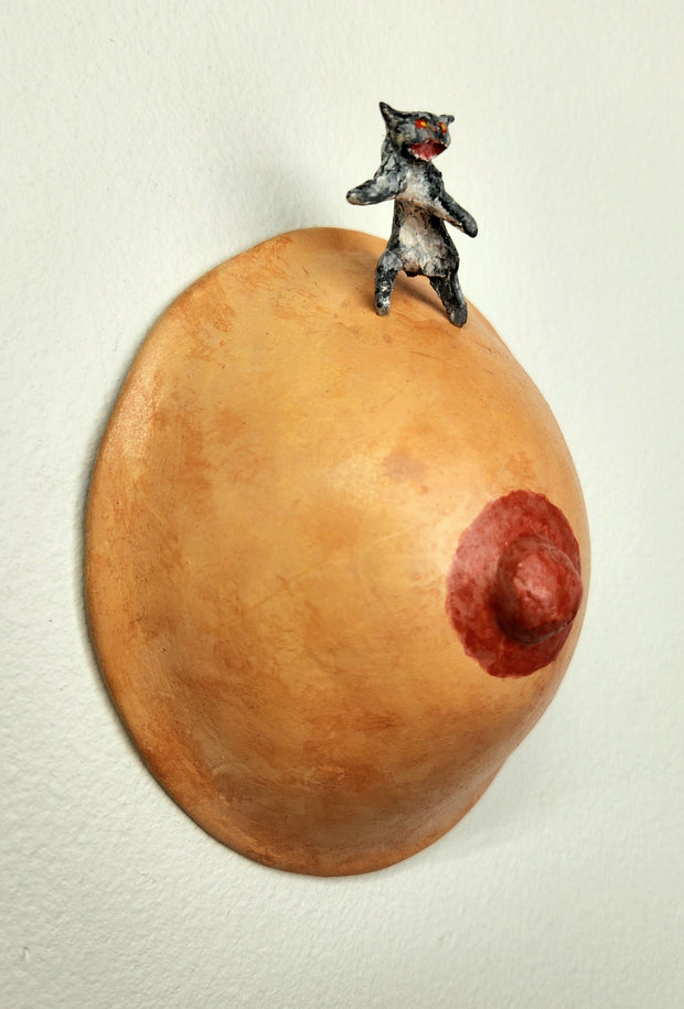Sculpture of a single boob with a painted nipple. A small sculpted gray tabby cat stands atop it on its hind legs, with a fierce expression.