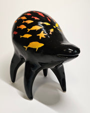 Black ceramic sculpture of a rounded body quadruped creature with an open mouth goofy smile. On its back are fish silhouettes in yellow, red and orange.