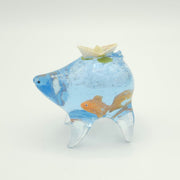 Resin sculpture of a blue rounded body quadruped creature with the illusion of water inside of its body. A goldfish swims at the bottom of its stomach and atop its back is a paper boat.