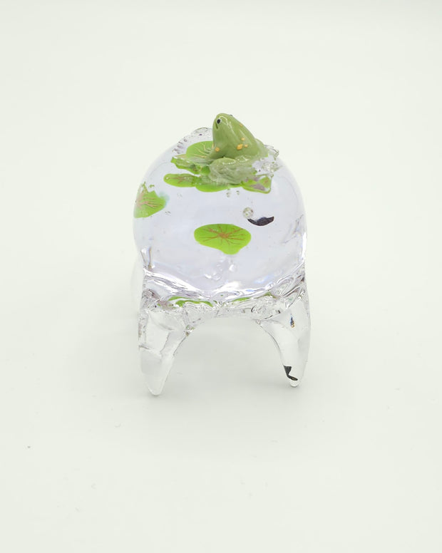 Resin sculpture of a clear rounded body quadruped creature with the illusion of water inside of its body. Green lily pads float within its body and atop its back is a green frog.