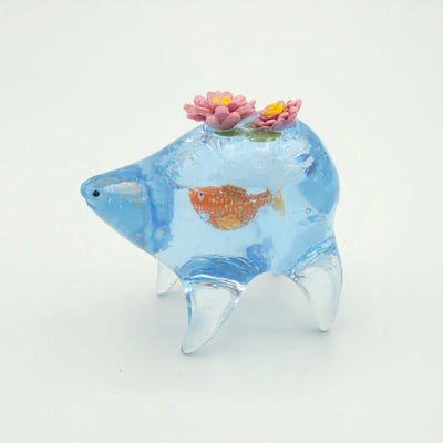 Resin sculpture of a blue rounded body quadruped creature with the illusion of water inside of its body. A large goldfish swims within its stomach and atop its back are 2 pink water lilies.