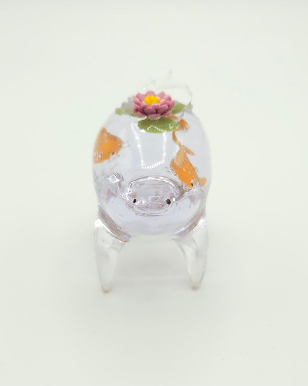 Resin sculpture of a clear rounded body quadruped creature with the illusion of water inside of its body. Several goldfish swim within its stomach and atop its back is a pink water lily with lily pads.