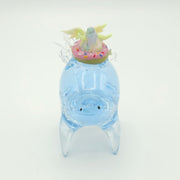 Resin sculpture of a blue rounded body quadruped creature with the illusion of water inside of its body. On its back is a pastel winged chubby creature in a donut shaped inflatable tube.