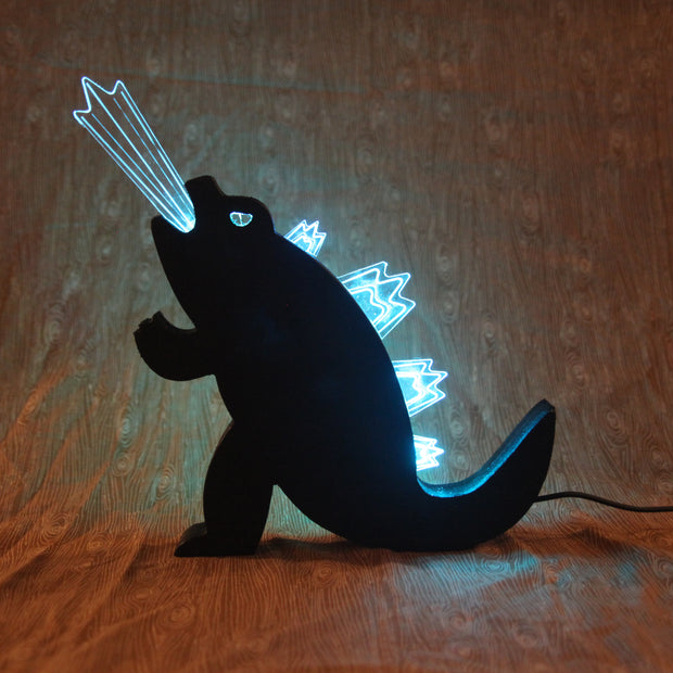 Die cut wooden silhouette of Godzilla, made into a lamp. A beam of light comes out of its mouth and it has lit up spikes along its back.