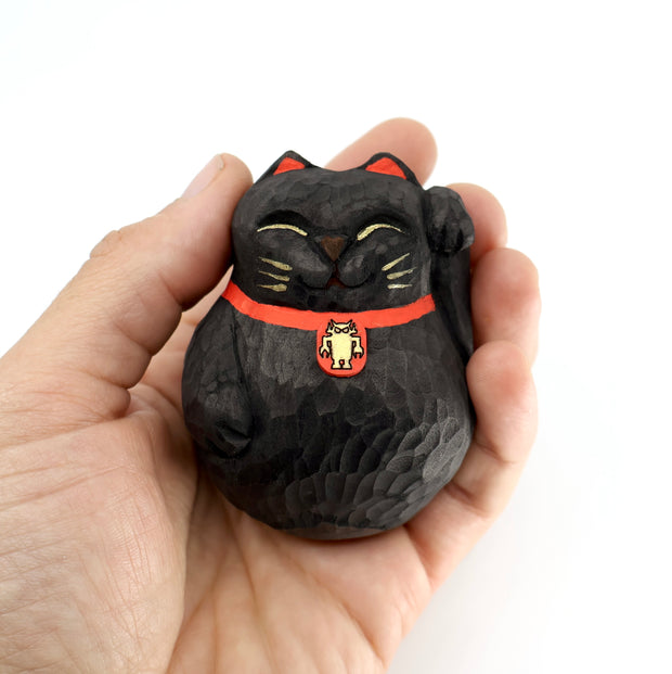 A rounded whittled wooden sculpture of a black maneki cats, smiling with closed eyes and one paw raised. It has gold whiskers and eyelids and wears a red collar with a gold Big Boss Robot on it. Held in someone's hand.