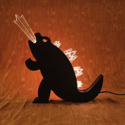 Die cut wooden silhouette of Godzilla, made into a lamp. A beam of light comes out of its mouth and it has lit up spikes along its back. 
