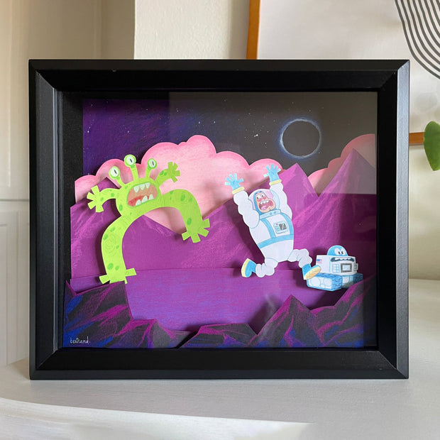 Framed paper cutting of a illustrative style astronaut and small robot running away comically from a green alien, who appears also startled. Space background is purple and pink.