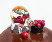 Clear resin sculpture of a quadruped creature with minimal features, and a rotund body. Inside its body are many small fruits with comical faces. It stands next to a basket of apples with similar comical faces. Both are on a brown glitter circular base.