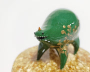 Small sculpture of a green quadruped creature with a small face and very rotund body. It has a single tiny gold horn and gold painted patterns along its side. It stands on a gold speckled clear base.