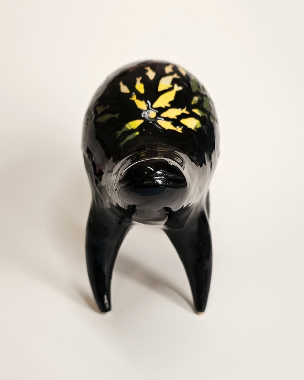 Black ceramic sculpture of a rounded body quadruped creature with an open mouth goofy smile. On its back are fish silhouettes in yellow and olive green.