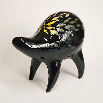 Black ceramic sculpture of a rounded body quadruped creature with an open mouth goofy smile. On its back are fish silhouettes in yellow and olive green.