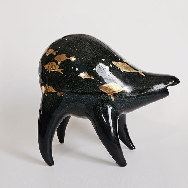 Black ceramic sculpture of a rounded body quadruped creature with an open mouth goofy smile. It has golden fish silhouettes swimming on its back, with gold splatters as well.