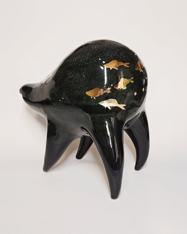 Black ceramic sculpture of a rounded body quadruped creature with an open mouth goofy smile. It has golden fish silhouettes swimming on its back, with gold splatters as well.