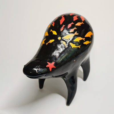 Black ceramic sculpture of a rounded body quadruped creature with an open mouth goofy smile. On its back are fish silhouettes in yellow, red and orange.