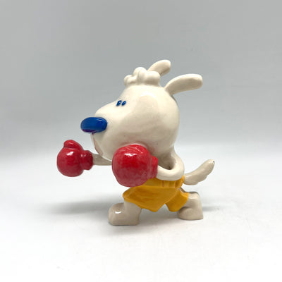 Ceramic sculpture of a small white cartoon dog, dressed like a boxer in yellow shorts and red boxing gloves. It stands in a punching position, with one hand extended out.