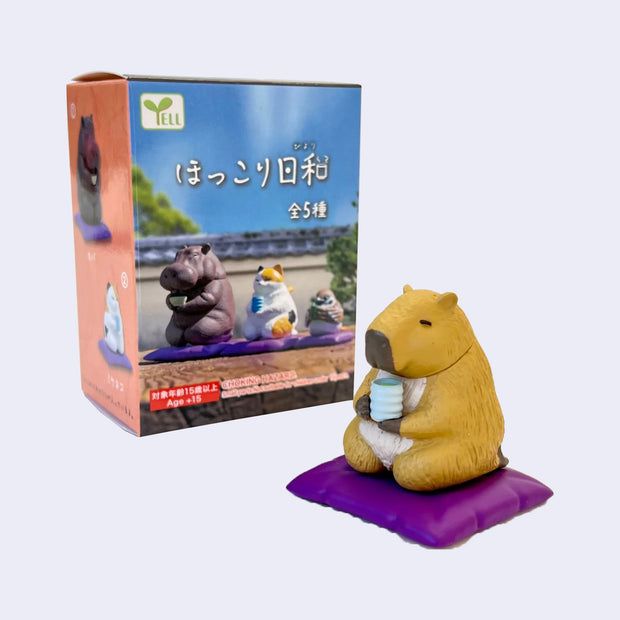 Small plastic figure of a capybara, sitting on a purple floor pillow and holding a ribbed cup of tea in its hands. Its eyes are closed peacefully, as it sits right in front of its product packaging.