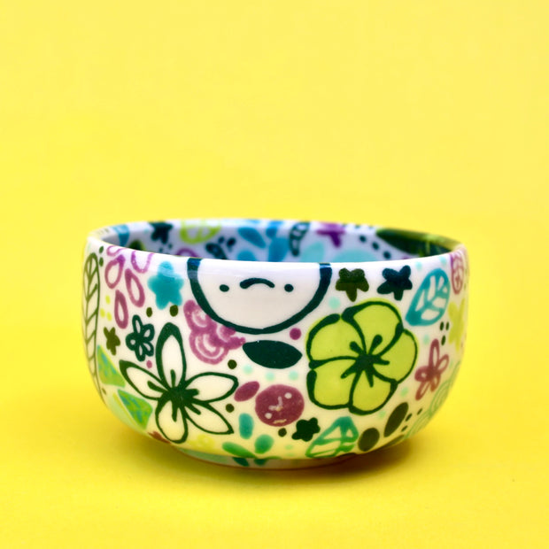 Small ceramic bowl, white with many paintings of floral patterns and fruits in cool tones such as green, blue and purple.