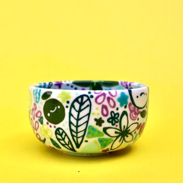 Small ceramic bowl, white with many paintings of floral patterns and fruits in cool tones such as green, blue and purple.