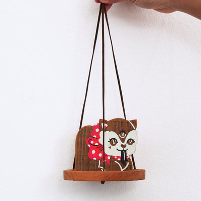 Wooden sculpture of a cat with 3 eyes, a red polka dot bow around its neck and a forked tongue. It sits on a round tray and hangs from 3 suede strings.