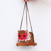 Wooden sculpture of a cat with a red masquerade style mask on, a white and pink polka dot bow and a smile. It sits on a round tray and hangs from 3 suede strings.