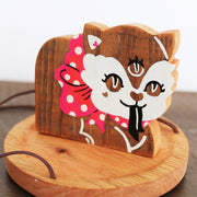 Wooden sculpture of a cat with 3 eyes, a red polka dot bow around its neck and a forked tongue. It sits on a round tray and hangs from 3 suede strings.