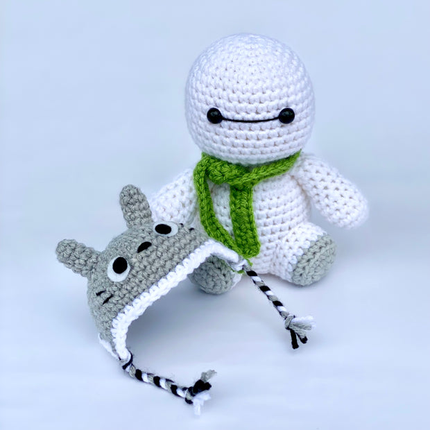 Crocheted version of Baymax from Big Hero 6, a white round edged robot with a kind face. It wears a green knit scarf and has a crocheted beanie nearby that resembles Totoro.