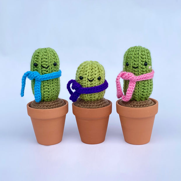 3 crocheted cacti in real clay pots, with crocheted soil and crocheted scarfs. Scarf colors are blue, purple or pink.