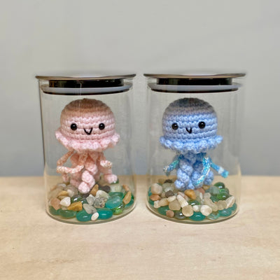 2 glass jars, each with a plush jellyfish inside with a cute face sewn on. At the bottom of each jar are colorful rocks. One jellyfish is pink and the other is blue.