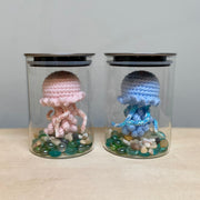 2 glass jars, each with a plush jellyfish inside with a cute face sewn on. At the bottom of each jar are colorful rocks. One jellyfish is pink and the other is blue.