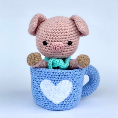 Crochet sculpture of a cute pink pig with an oversized head and a teal scarf around its neck sitting inside of a blue coffee mug. The mug is also crocheted and has a white heart as the center design.