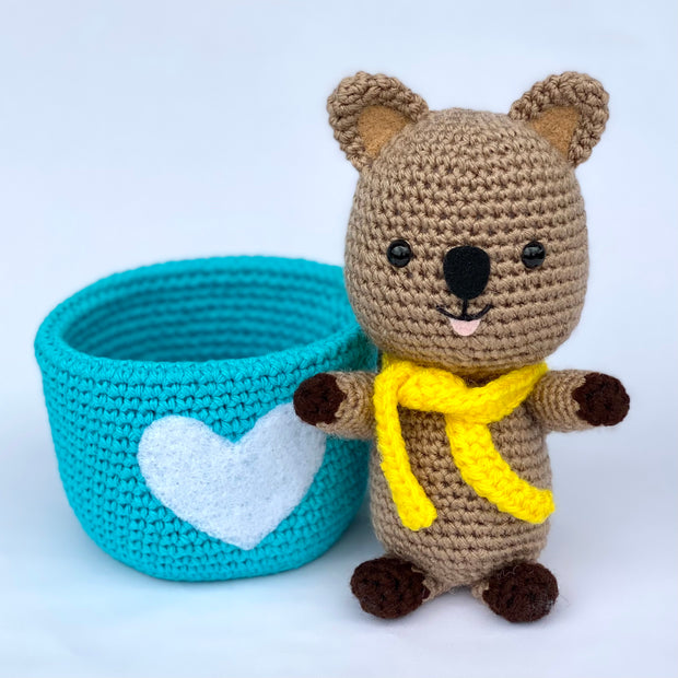  Crochet sculpture of a quokka, a small brown marsupial, wearing a yellow knit scarf and sitting outside of a blue crocheted mug with a white heart as the center design.