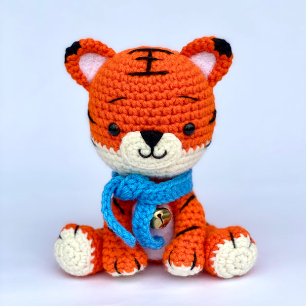 Crocheted tiger with a slightly oversized head and a sweet, smiling expression. It sits and wears a blue knit scarf with a metal bell around its neck.
