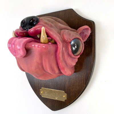 Sculpture of a pink dog's head with a large underbite and a shiny nose and large eyes. Its tongue sticks out and it's head is mounted to a wooden plaque, like a commemorative deer head.