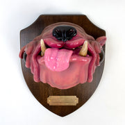 Sculpture of a pink dog's head with a large underbite and a shiny nose and large eyes. Its tongue sticks out and it's head is mounted to a wooden plaque, like a commemorative deer head.