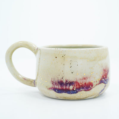 Short cream colored ceramic mug with a large, thin handle. Abstract subtle red and purple coloring is at the bottom with gold speckles.