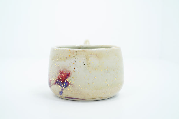 Short cream colored ceramic mug with a large, thin handle. Abstract subtle red and purple coloring is at the bottom with gold speckles.