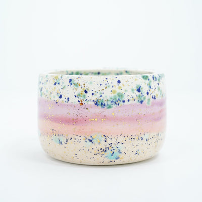 Short ceramic bowl, mostly off white with a pink stripe running through it and blue speckles all over. Gold specks are also on the surface.