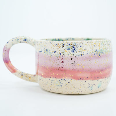 Short ceramic mug with thin large handle, mostly off white with a pink stripe running through it and blue speckles all over. Gold specks are also on the surface.