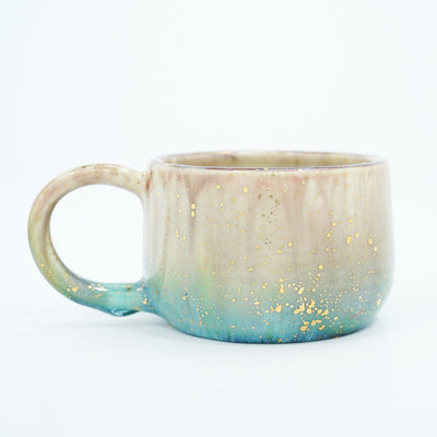 Small ceramic mug with large, thin handle and with a gradient of light brown to blue and gold speckles.