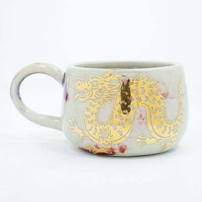 Ceramic cup with a thin handle, off white with subtle red splatter coloring. A gold dragon is painted onto the front of the mug.
