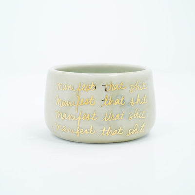 Short ceramic bowl, off white with four lines of repeated text that reads "manifest that shit" in gold cursive.