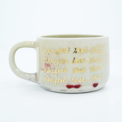 Short ceramic mug, off white with four lines of repeated text that reads "manifest that shit" in gold cursive.
