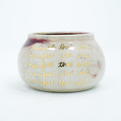 Short ceramic pot, off white with four lines of repeated text that reads "manifest that shit" in gold cursive.
